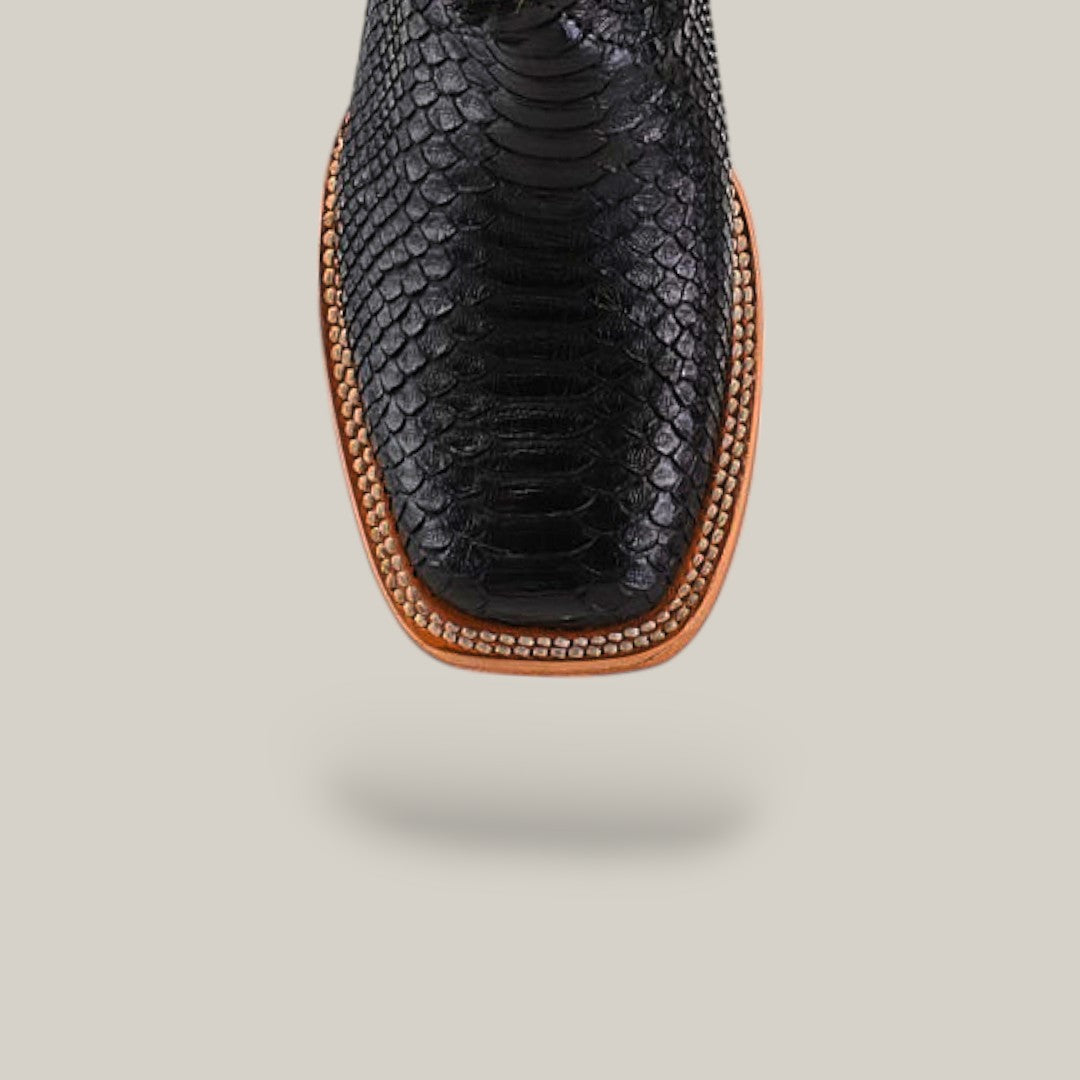 Close-up of Exotic Python Black - Square Toe - Kids Boots featuring a snakeskin texture, tan leather trim, and detailed white stitching on the edge, displayed on a plain beige background.