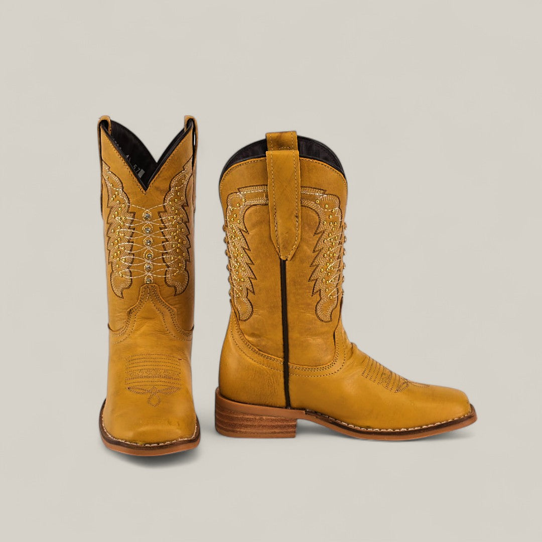 The Orion Yellow - Square Toe Kids Boots are tan with decorative stitching, wooden heels, pull straps, and a stylish square toe. Shown on a plain light background, one boot stands upright while the other tilts forward.