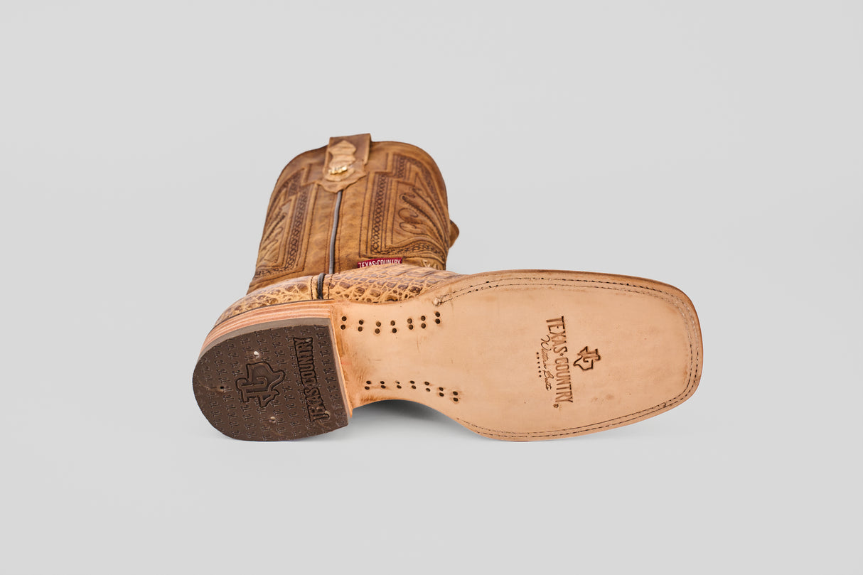 The Exotic Caiman Belly - Antique - Square Toe boot is shown on its side, highlighting the detailed shaft embroidery and stamped TEXAS COUNTRY logo on the tan sole. The square toe design features a black rubber heel grip with a star motif.