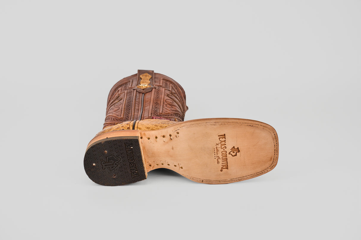 The Exotic Caiman Belly - Camel - Square Toe boot is shown on its side, revealing the engraved leather sole with TEXAS COUNTRY and a star logo, detailed brown stitching, and a square toe on a plain light background.