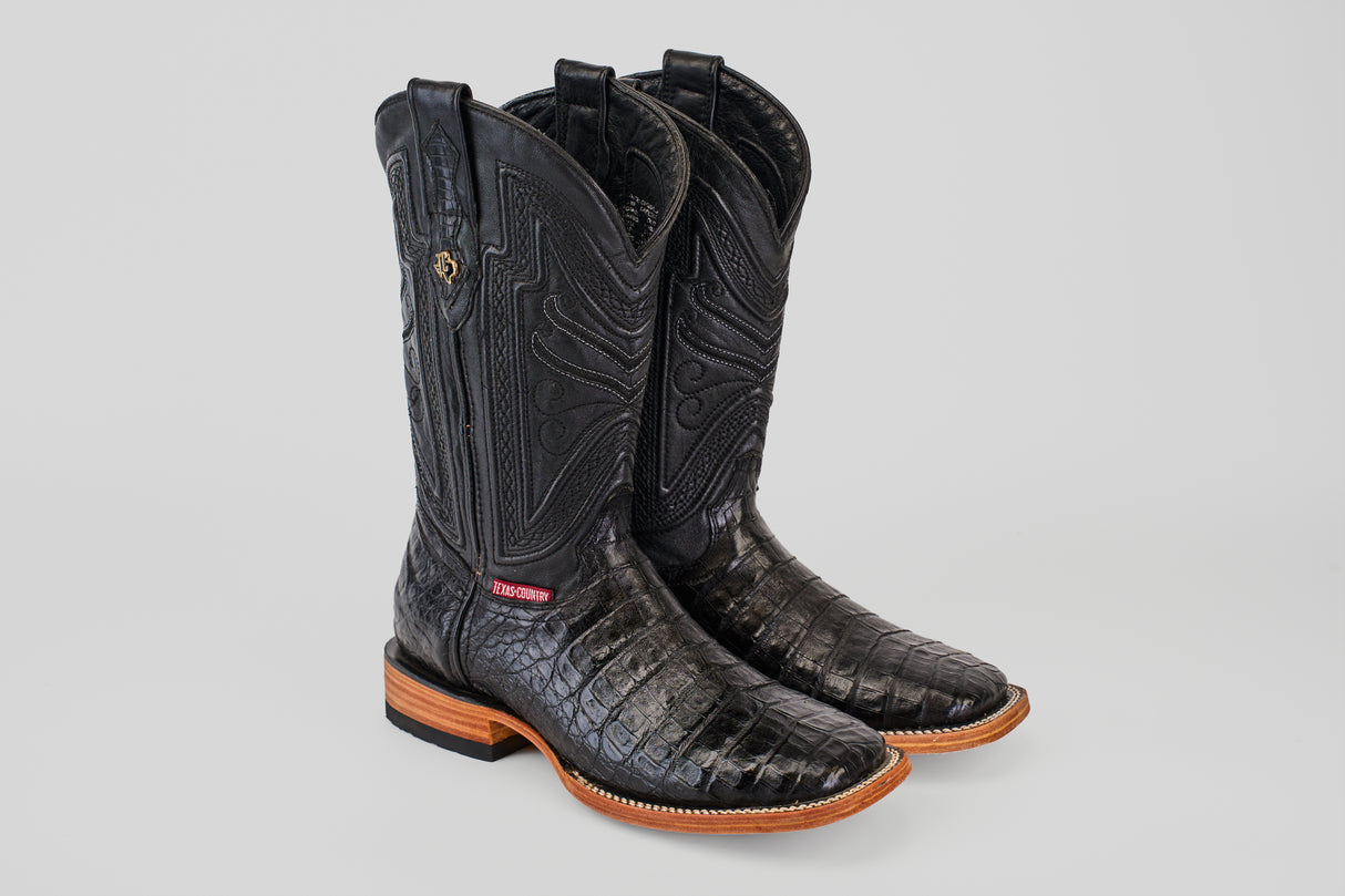 The Exotic Caiman Belly - Black - Square Toe boots feature embossed patterns, wooden soles, and a red and white side tag, showcased against a plain light background.