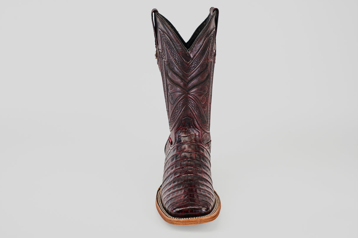 The Exotic Caiman Belly - Black Cherry cowboy boot is shown upright and front-facing, highlighting its detailed stitching, unique black cherry finish, square toe, and sleek design against a plain grey background.
