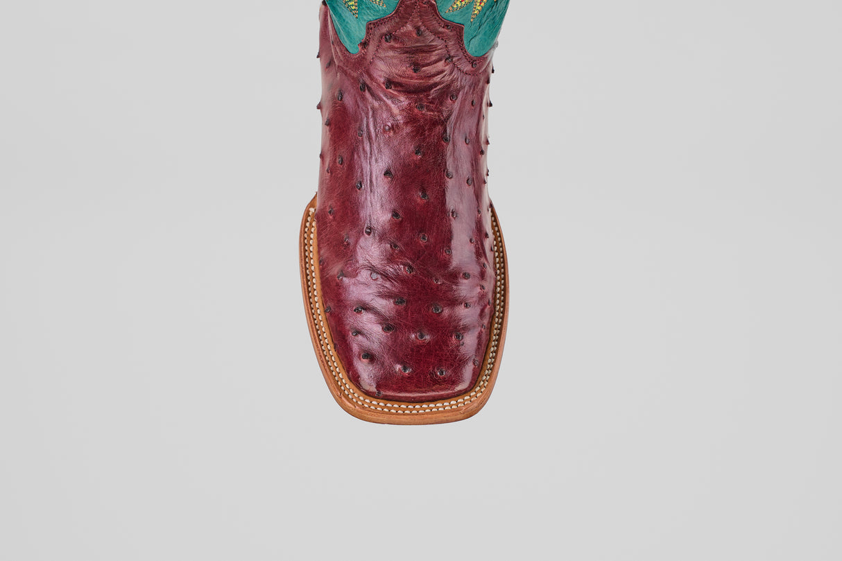 A close-up of the Rusty - Wine - Square Toe boot shows its rich ostrich leather texture, tan stitching, and turquoise detailing at the top.