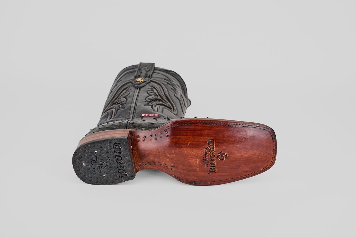 The Exotic Ostrich - Black - Rodeo Toe boot rests on its side, displaying its detailed brown leather sole with Tecovas branding and decorative stitching—an elegant piece of western wear.