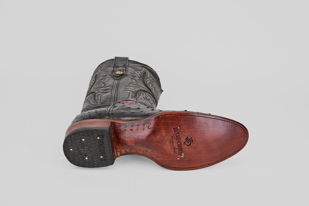 A single Exotic Full Quilt Ostrich - Black - Round Toe cowboy boot lies on its side, highlighting intricate upper stitching, smooth brown leather sole, visible branding, and metal tacks on the heel.