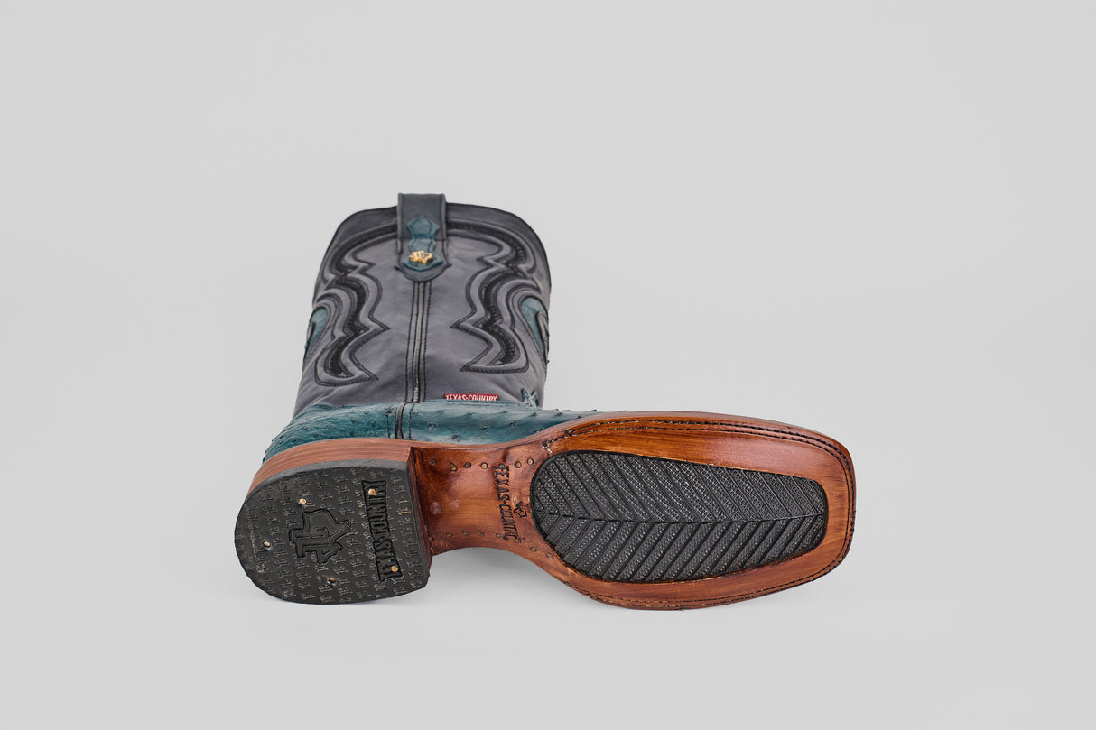 The Austin - Gasoline Green - Square Toe men's western boots feature black, teal, and brown leather. One boot stands upright to display its decorative shaft; the other lies sideways, revealing a textured sole with rubber grip and detailed stitching.