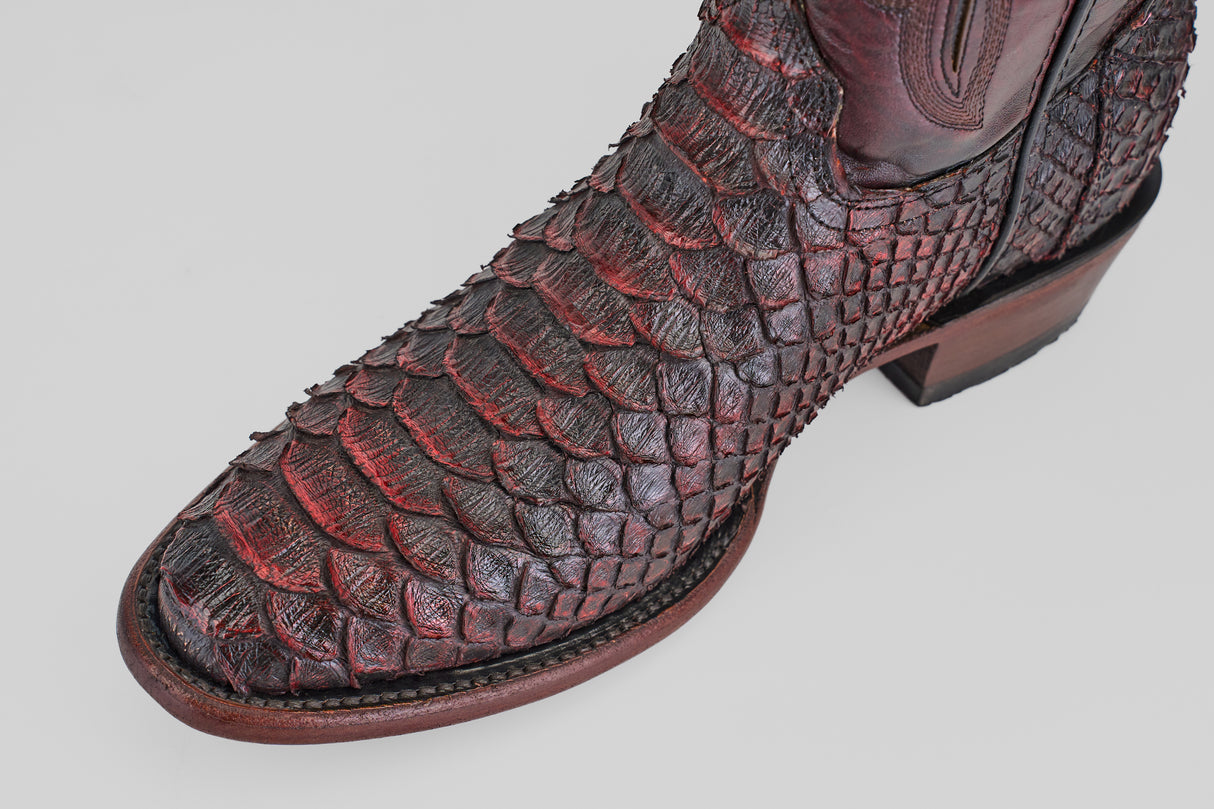 A close-up of the Tanner Jumbo - Black Cherry - Round Toe cowboy boot, featuring reddish-brown, textured exotic leather with scale-like detail, intricate stitching, and a sturdy wooden heel.