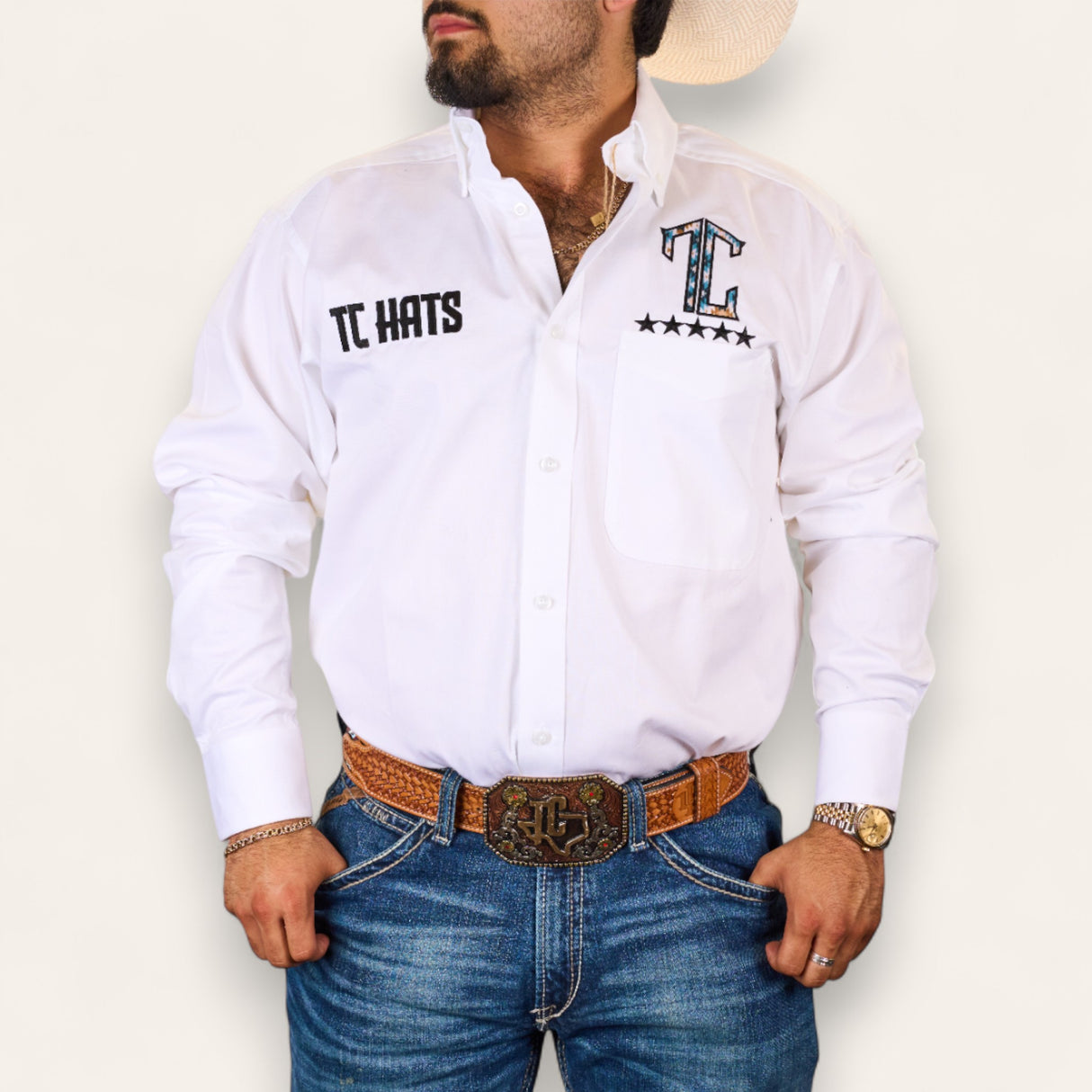 A man stands with hands on hips against a plain background, wearing the TC Hats - White Blue Aztec shirt made from lightweight fabric, blue jeans, a brown woven belt with a large buckle, wristwatch, and a light cowboy hat.
