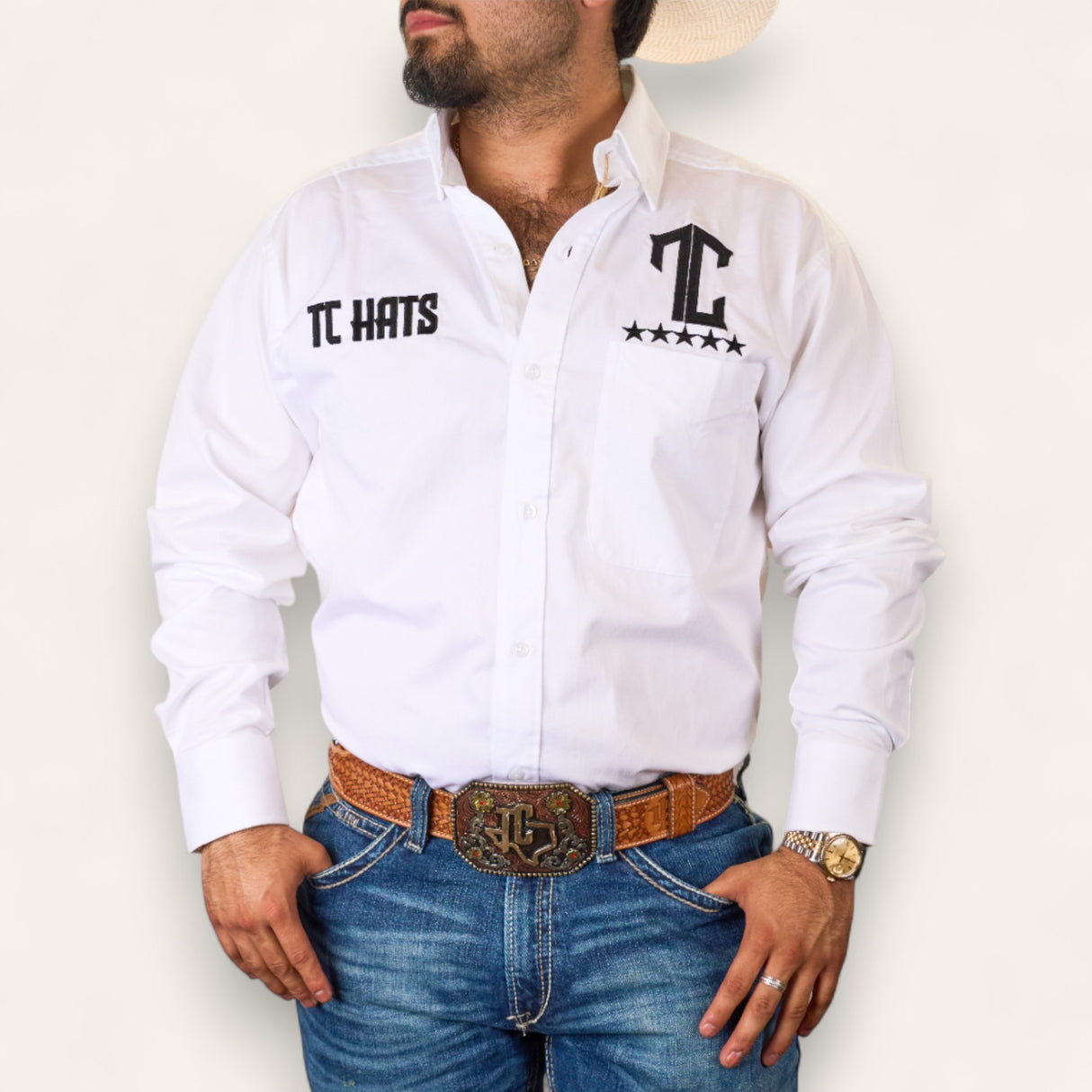 A man in a premium white button-up shirt with TC Hats - White & Black star designs, blue jeans, a brown belt with a large buckle, a watch, and a light cowboy hat stands against a plain background.