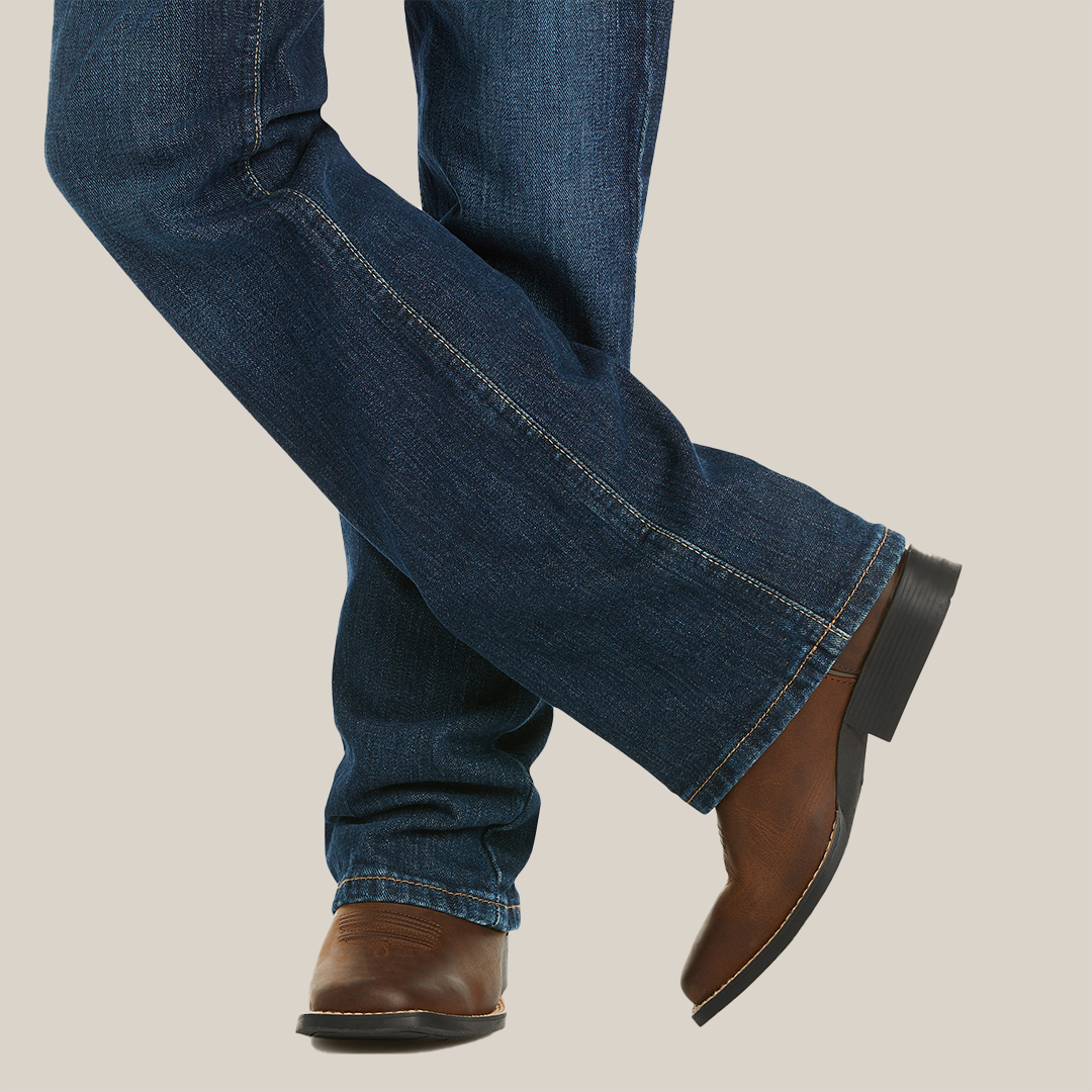 A person is casually standing with legs crossed, showcasing Ariat Boys B4 Legacy Dark Wash Relaxed Fit Boot Cut Jeans, paired perfectly with brown leather cowboy boots featuring a black heel against a neutral beige backdrop.