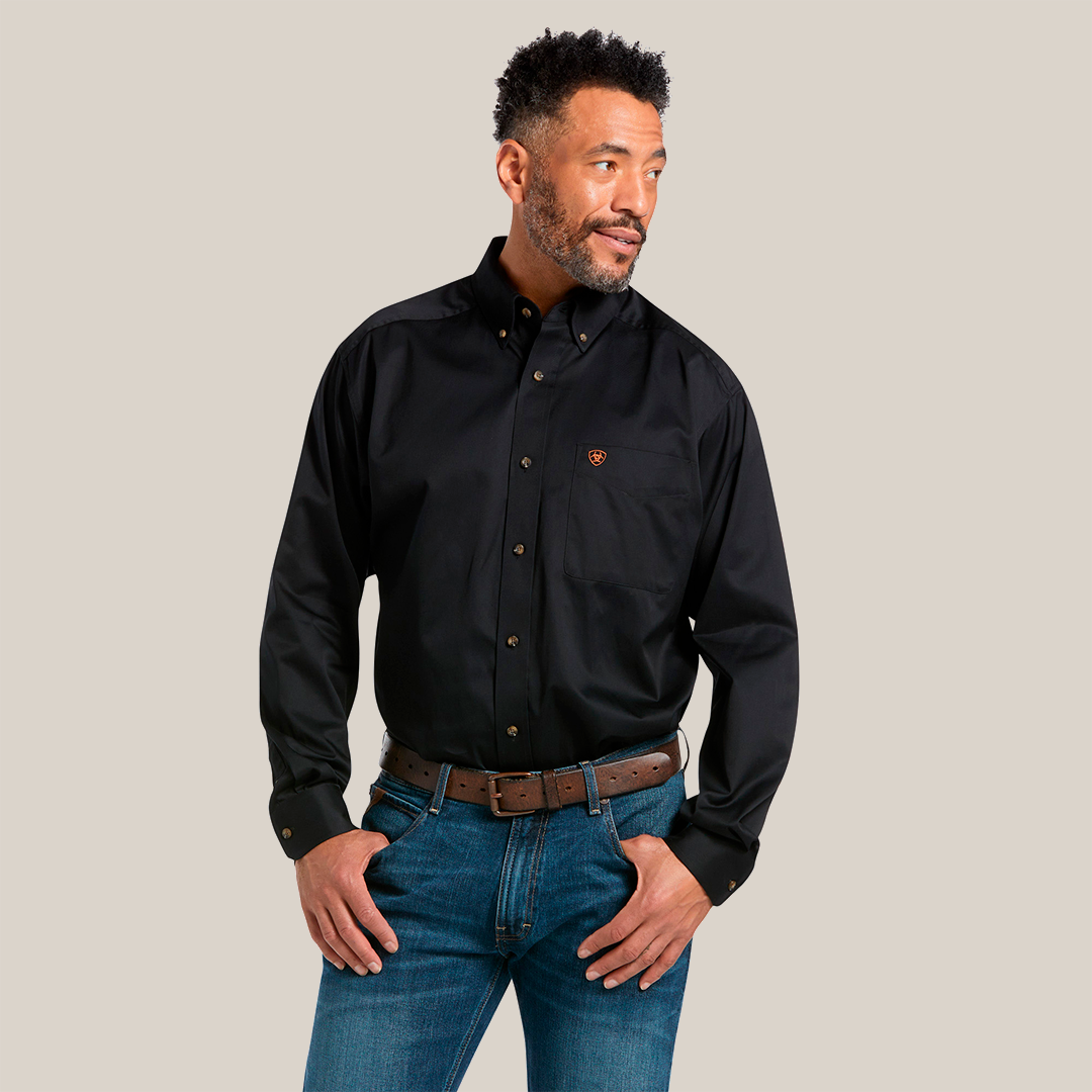 A man in a Solid Twill Classic Fit Shirt Black - 10000502, featuring embroidered chest pocket details, pairs it with blue jeans and a brown belt. Hands in pockets, he looks to his left.