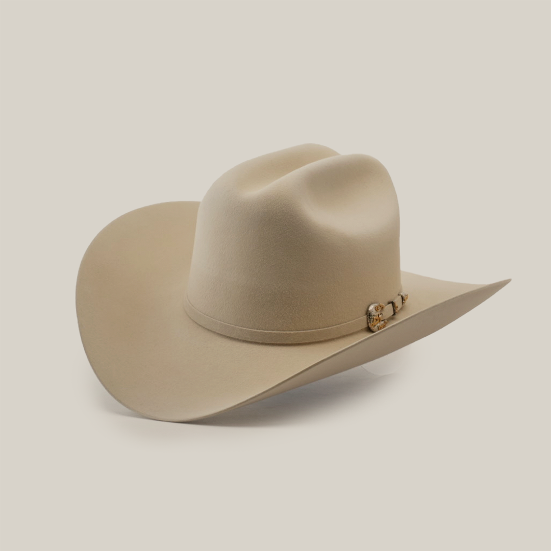 The 1000X Imperial Belly hat is a beige cowboy style with a wide brim, adorned with a decorative band featuring a small metallic emblem, set against a plain white background.