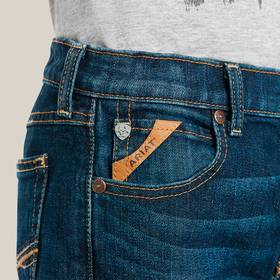 A person is wearing Ariat Boys B4 Legacy Dark Wash Relaxed Fit Boot Cut Jeans, featuring yellow stitching, a small front pocket with a brown leather Ariat label, and a silver button on the waistband. A gray shirt peeks out above the jeans.