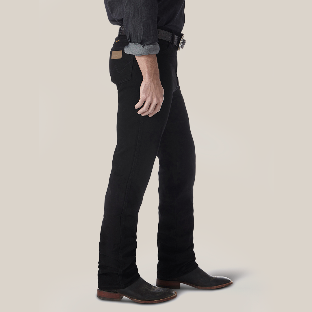 Side view of a person in Wrangler Cowboy Cut Slim Fit Jean - 0936WBK with a brown leather patch, black belt, and dark shirt with rolled sleeves. Complemented by black boots, the outfit stands out against a plain light background, highlighting the premium denim style.