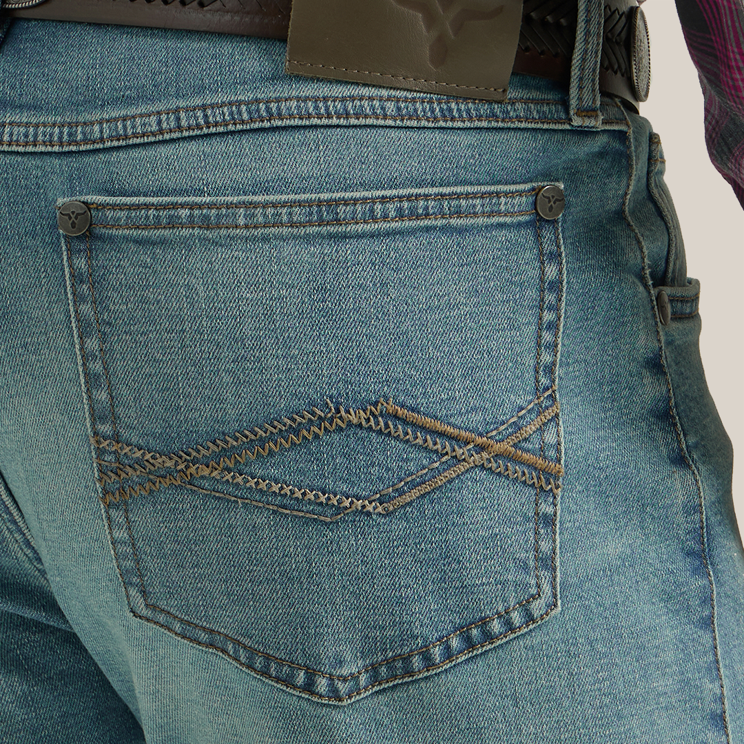 Someone in a plaid shirt, Wrangler 20X Mens Light Wash Shade Bootcut Stretch Jeans, and brown cowboy boots stands against a plain background. The light blue jeans are slightly faded and secured with a brown belt featuring an oval buckle.