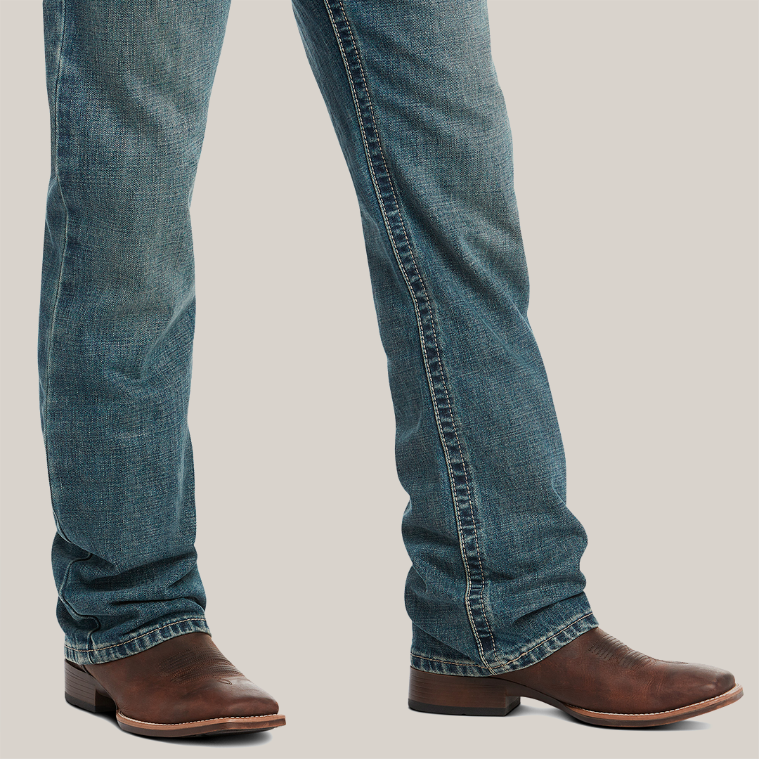 Wearing M4 Low Rise Boundary Boot Cut Jeans in Gulch and brown leather cowboy boots, a pair of legs is posed against a white backdrop. The slightly faded cotton denim features a relaxed fit with straight legs, while the boots boast low heels and rounded toes.