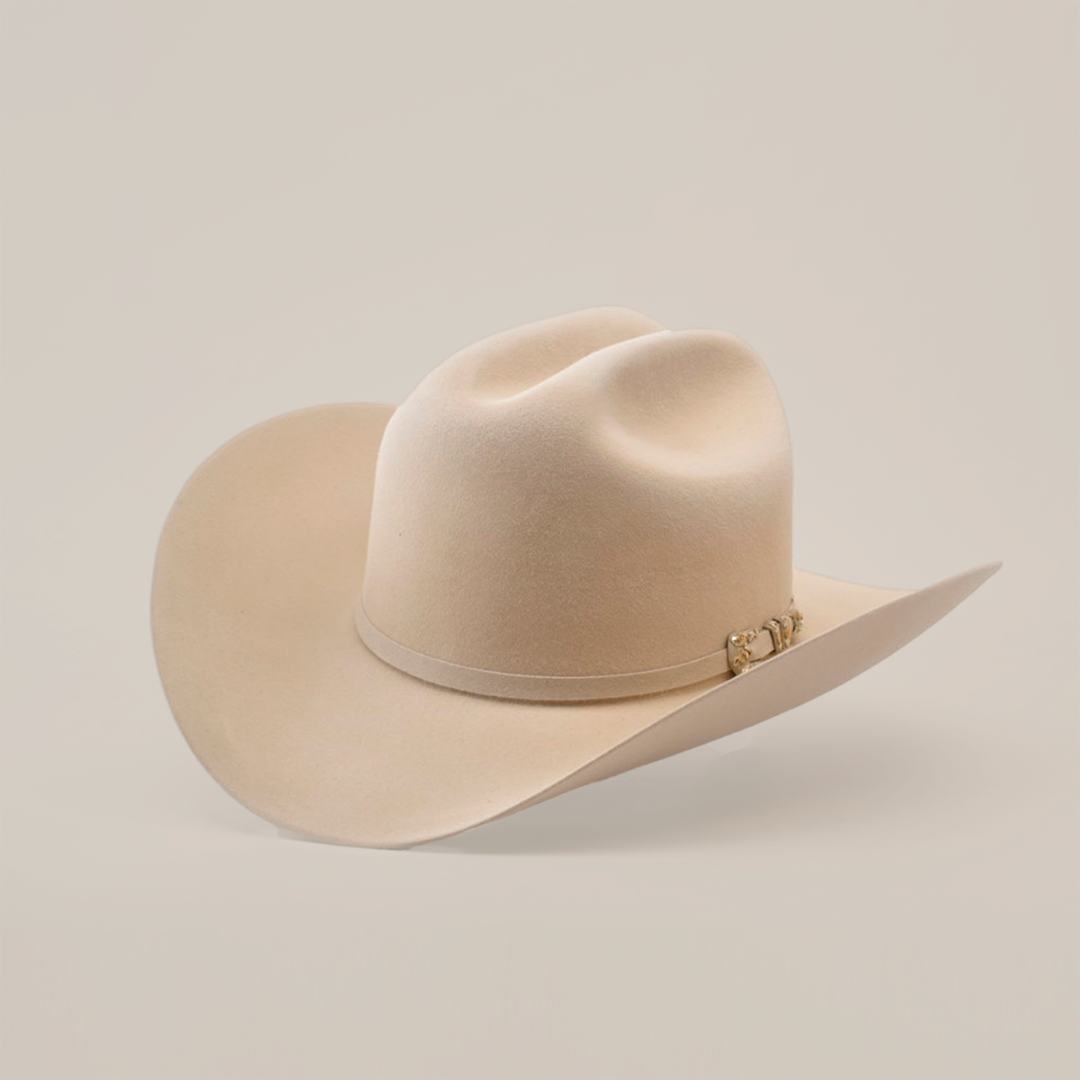 The 10X Jerarca Belly is a cream-colored cowboy hat made from luxurious fur felt, featuring a wide brim and distinctive crown crease. Adorned with a decorative metallic embellished band, this classic western headwear evokes Larry Mahans timeless style against a white backdrop.