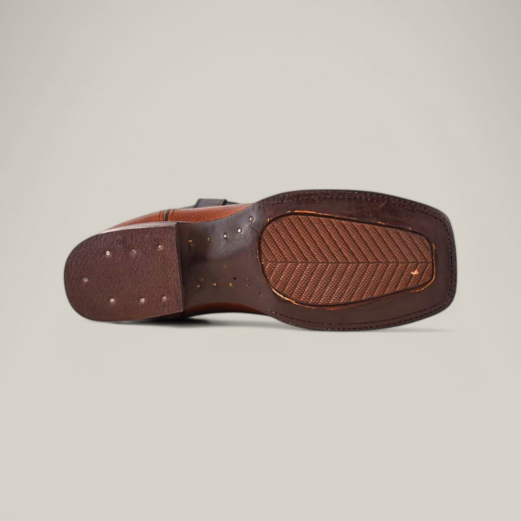 The image features the sole of a Grisly Tan - Square Toe boot, made of premium leather with stitching and small perforations. This Western-style shoe has a textured grip and rectangular heel, set against a plain light gray background.