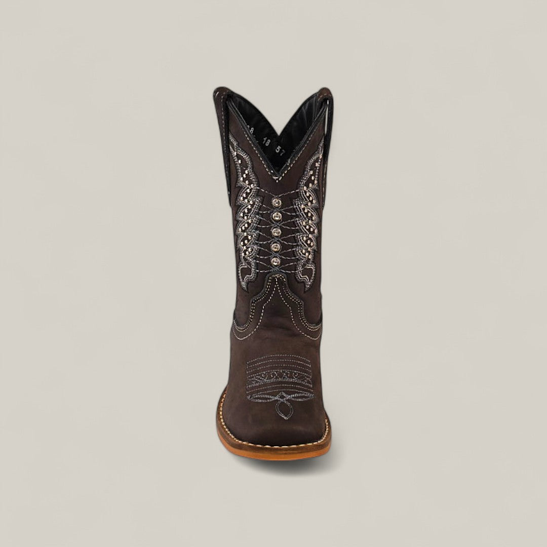 The Orion Suede Oxford - Square Toe Kids Boot is shown from the front in brown suede, featuring intricate white and silver stitching with decorative patterns, set against a plain light gray background.