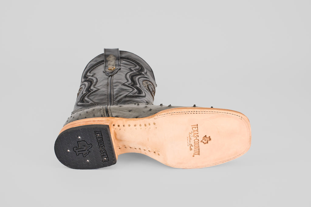 A single Exotic Full Quilt Ostrich - Gray - Square Toe boot lies on its side, showing its tan leather sole with visible stitching, Texas Country logo, black rubber heel, and classic ostrich-inspired decorative stitching on the upper.