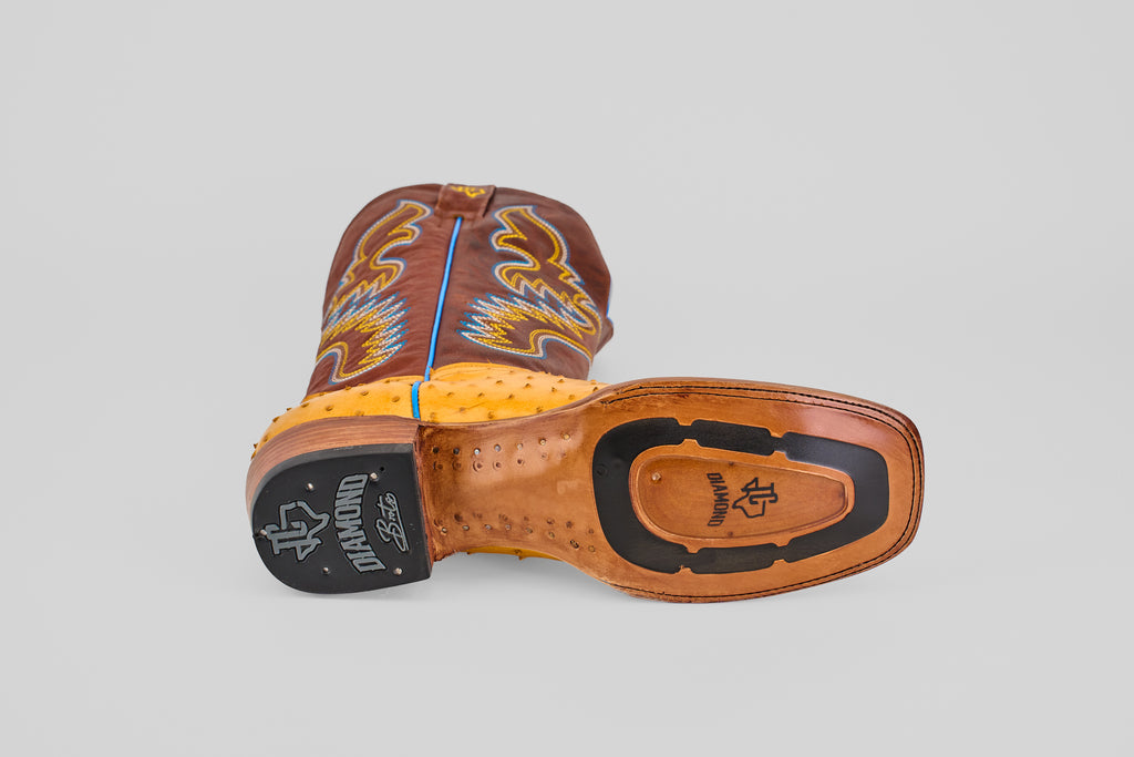 The Ransom - Buttercup - Square Toe boot is shown on its side, revealing the sole's detailed stitching, rubber heel, and brand logo. Blue and yellow embroidery decorates this Platinum Collection design.