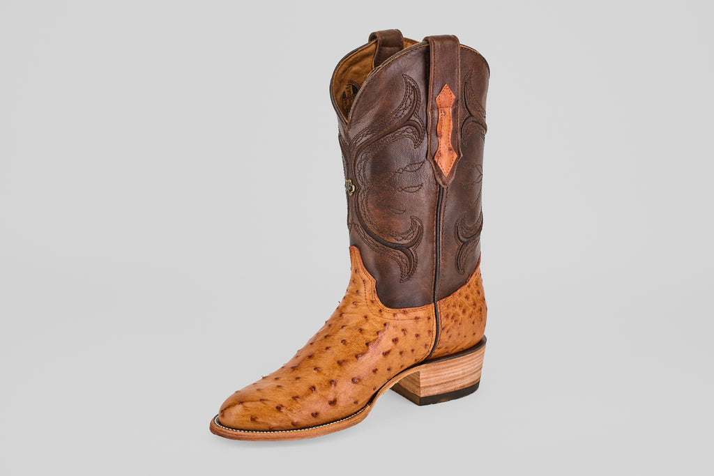 The Austin - Brandy - Roper Toe is a single men's cowboy boot with a tan ostrich leather foot, dark brown leather shaft, decorative stitching, and a stacked wooden heel, displayed on a plain gray background.