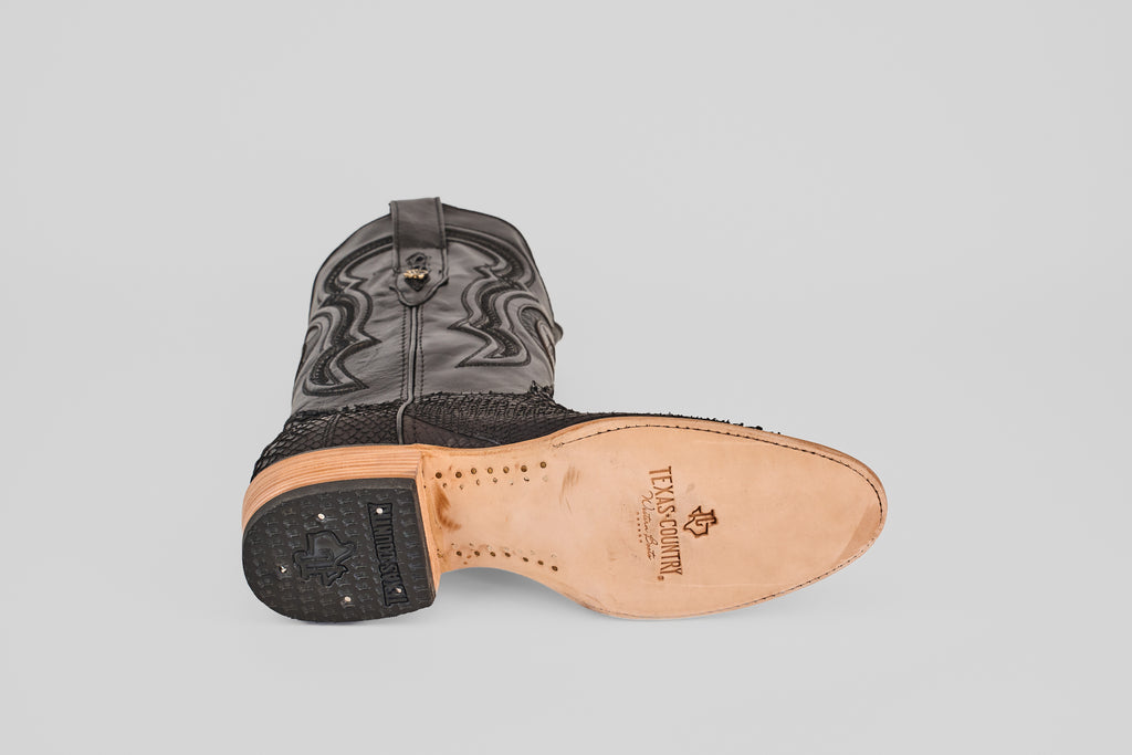 The Tanner – Matte Black – Roper Toe boot is shown on its side, highlighting the “Texas Country” stamped leather sole. This western-inspired boot features textured matte black leather and decorative stitching on the shaft.