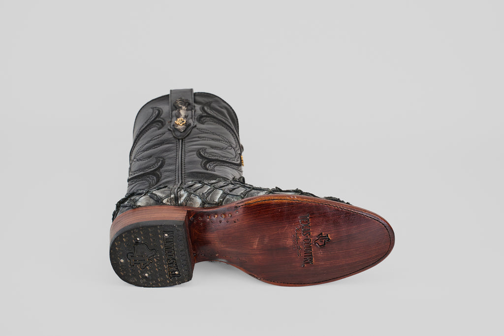 The Conor - Newspaper - Roper Toe boot in black exotic leather lies on its side, displaying the worn sole with branding and a textured heel against a light gray background—a classic for western boot enthusiasts.