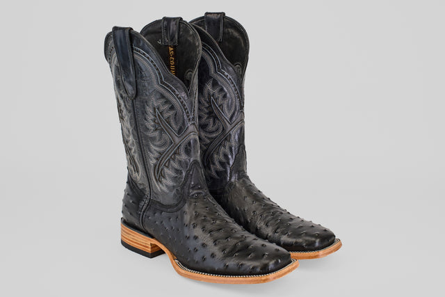 The Ostrich Black - Embossed Leather - Square Toe (E424) boots feature intricate stitching, textured patterns, a light brown wooden sole, and low heels—classic western style—displayed on a plain gray background.