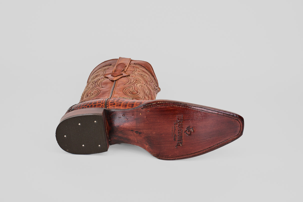 The Lizard Cogñac Embossed Leather Versy Toe (E201) cowboy boot is shown on its side, displaying its textured sole with metal studs at the heel and engraved text, plus decorative stitching that highlights classic western craftsmanship.