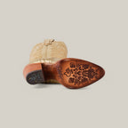 A single beige Linda Tall Shaft Gold - J Toe cowboy boot rests sideways, revealing an intricately engraved brown sole adorned with floral designs and embossed logos. As part of the Platinum Collection, its decorative stitching enhances its classic western style.