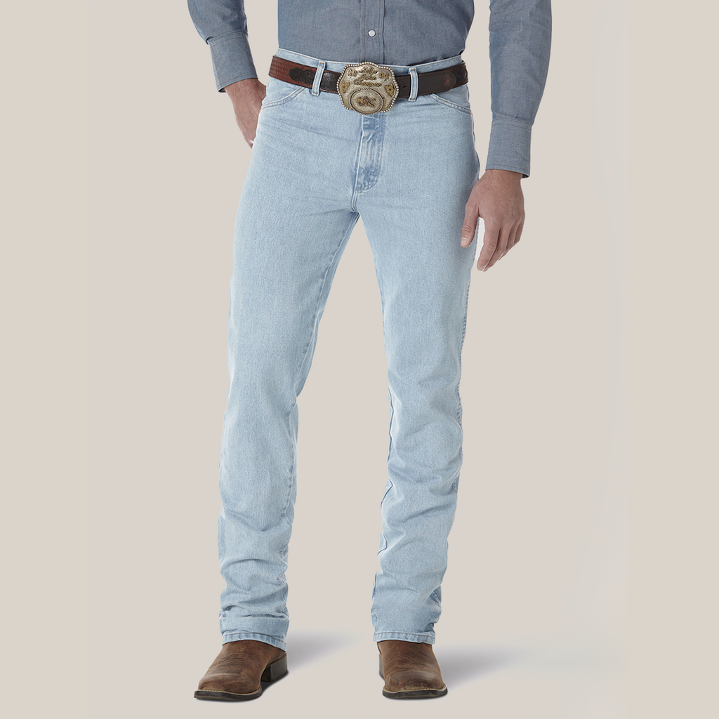 A person in Wrangler Cowboy Cut Slim Fit Jeans, wearing a brown belt with an ornate buckle, blue shirt, and brown boots. The premium denim fits perfectly against a plain light background, although their head is not visible.